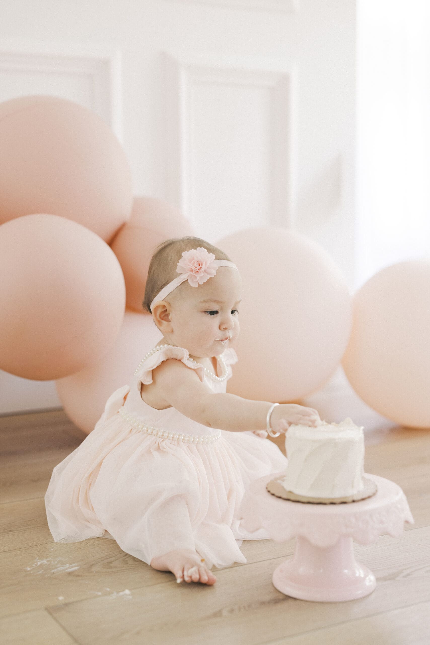 Baby girl smiling during a pink first birthday cake smash session with soft feminine decor, frosting covered hands, and playful milestone portraits in a Hampton Roads studio.